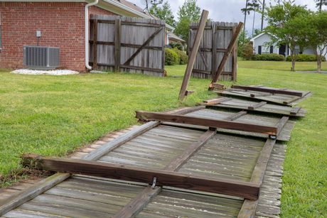 Fallen wooden fence panels lying along a grassy side yard beside a brick house and open gate.