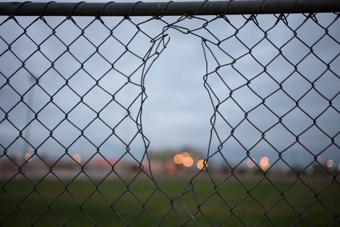 Broken chain-link fence with a torn opening, overlooking a blurred field and distant lights at dusk