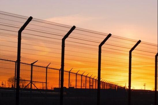 Sunset behind a chain-link fence and security posts, silhouetted against an orange sky