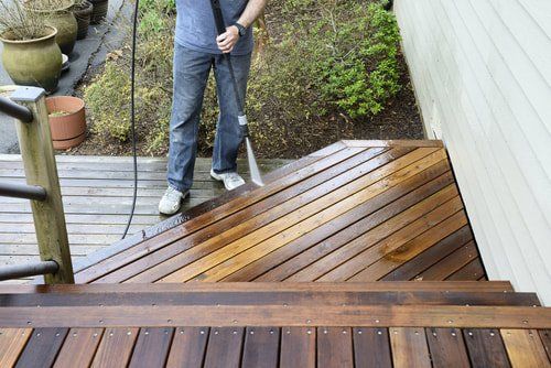 Person holding a hose sprays water on a wet wooden deck beside a house and garden.