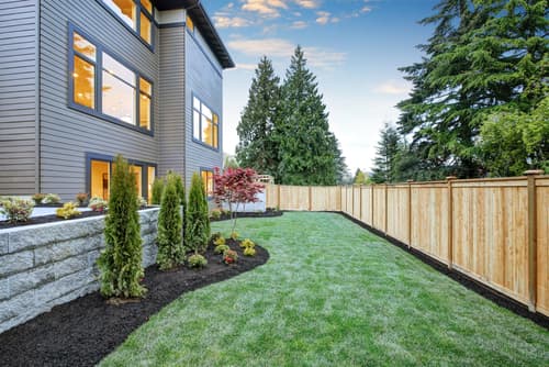 Modern house backyard with green lawn, wooden fence, and landscaped garden beds at dusk