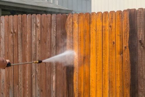 Person pressure-washing a wooden fence, revealing a bright cleaned section.