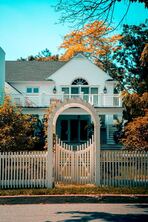 White house with a circular front porch and gate, framed by a white picket fence and autumn trees.