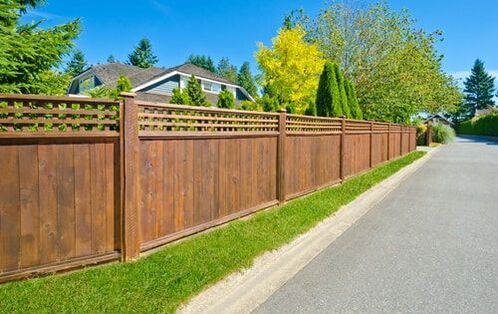 Wooden privacy fence along a quiet residential street with houses and green trees behind it.