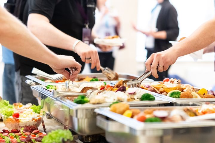 People use metal tongs to serve themselves from large buffet trays filled with various prepared dishes.