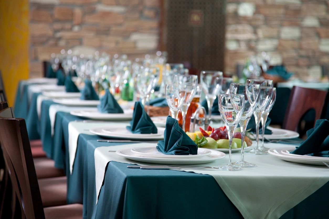 A long dining table set with teal tablecloths, folded napkins, and glassware in a rustic room with stone walls.