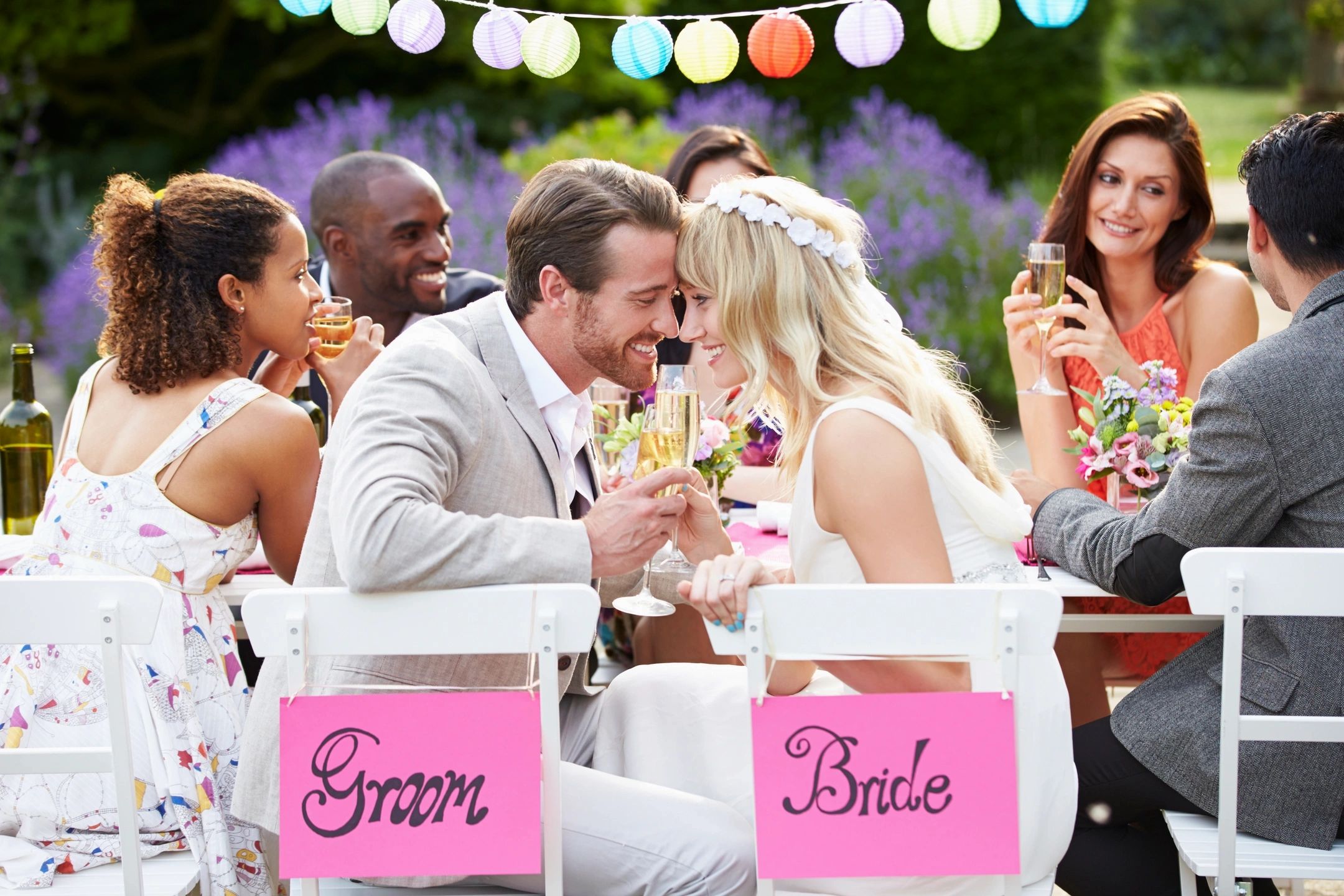 A couple toast with champagne at an outdoor wedding reception, with Groom and Bride signs on their chairs.