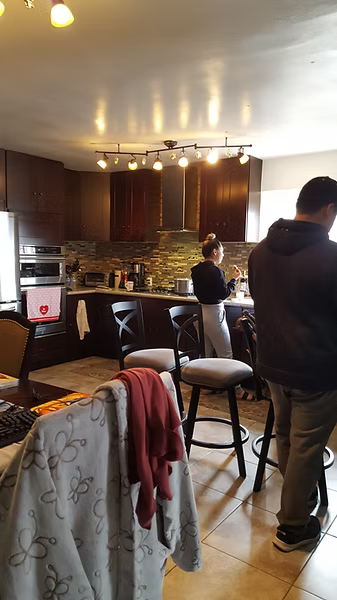 A person stands in a kitchen with dark cabinets and a tile backsplash while another person walks in the foreground.