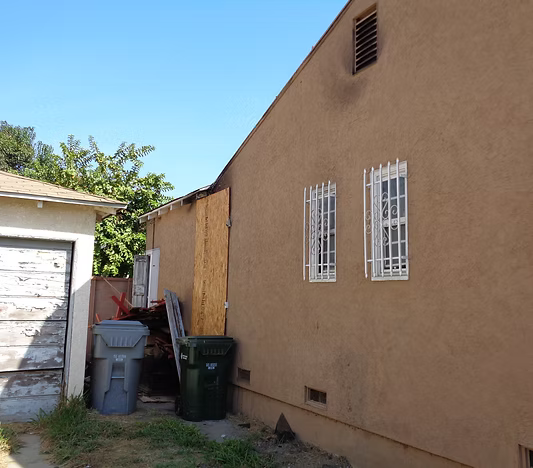 A tan stucco house exterior showing two windows with security bars, a boarded-up doorway, and trash bins in the yard.