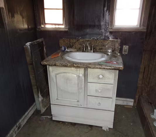 A white bathroom vanity with a granite countertop sits against a scorched, soot-covered wall beneath two small windows.
