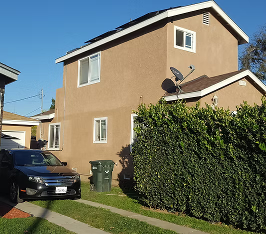 A two-story tan stucco house with solar panels on the roof, a driveway with a dark car, and a tall green hedge.