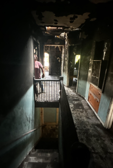 Damaged hallway with burnt walls and ceiling, leading to a bright doorway; person stands near the left railing.