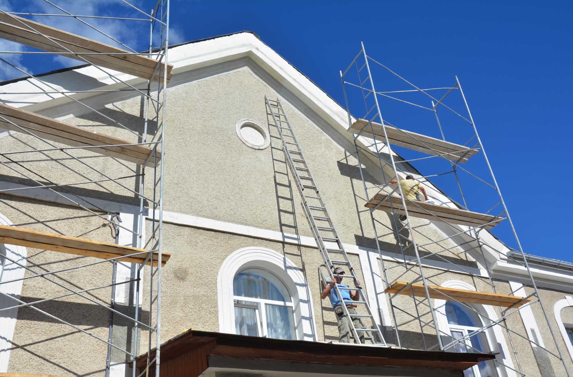 A worker on a ladder performs maintenance on the beige stucco exterior of a building under construction with scaffolding.
