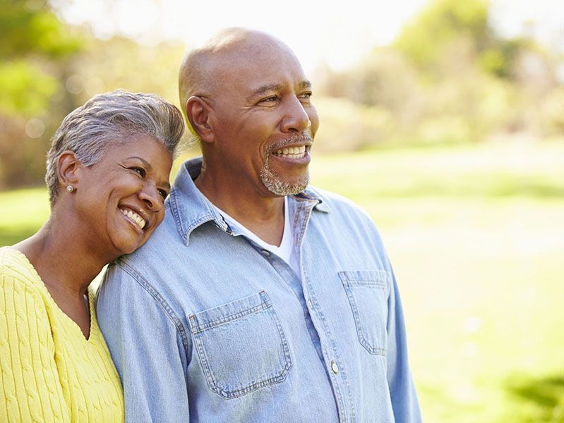 Two smiling adults standing close together outdoors in sunlight, wearing casual shirts.