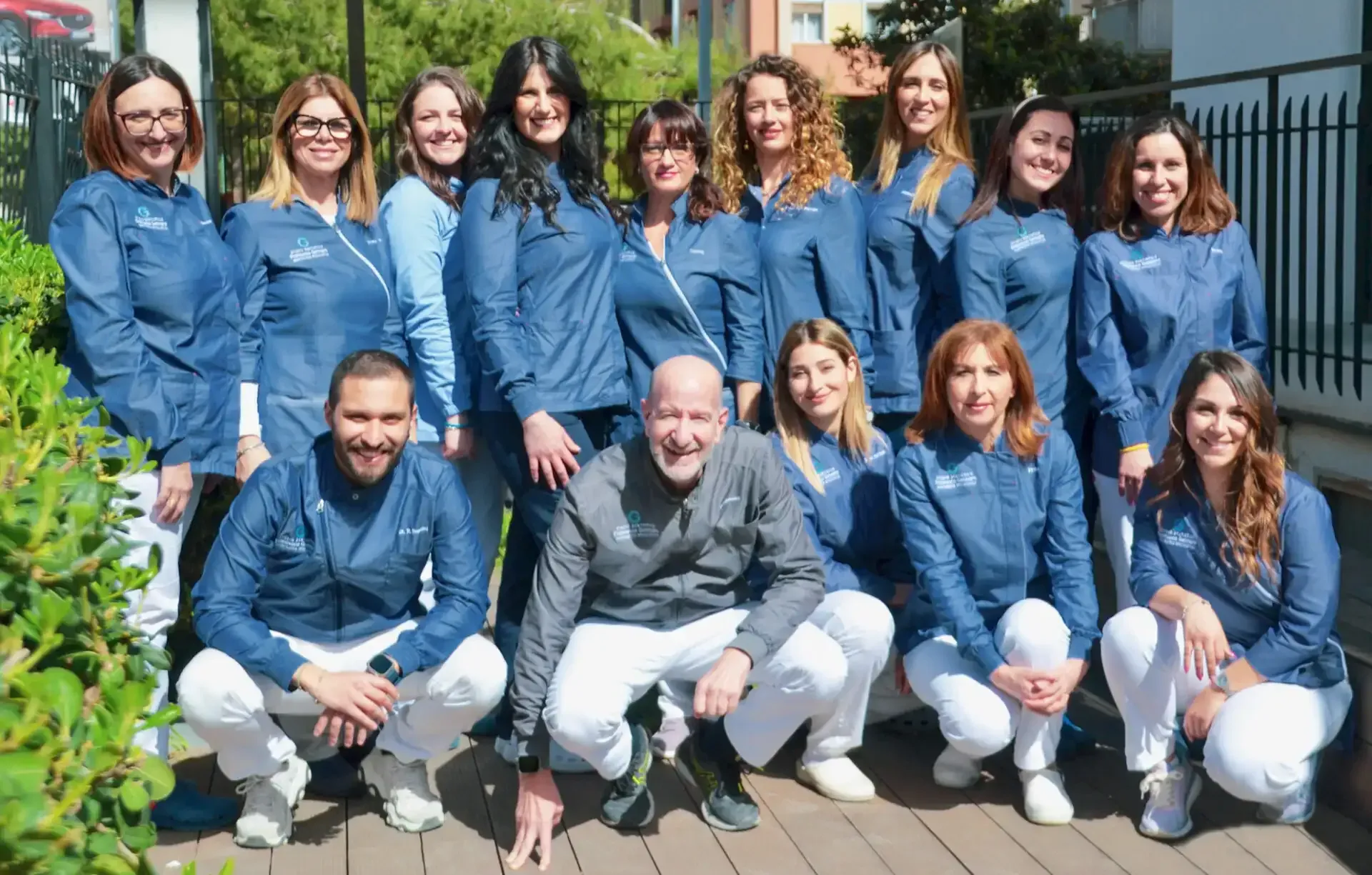 Gruppo di persone in posa all'aperto, con indosso camicie blu e pantaloni bianchi abbinati, sorridenti e disposte su due file.