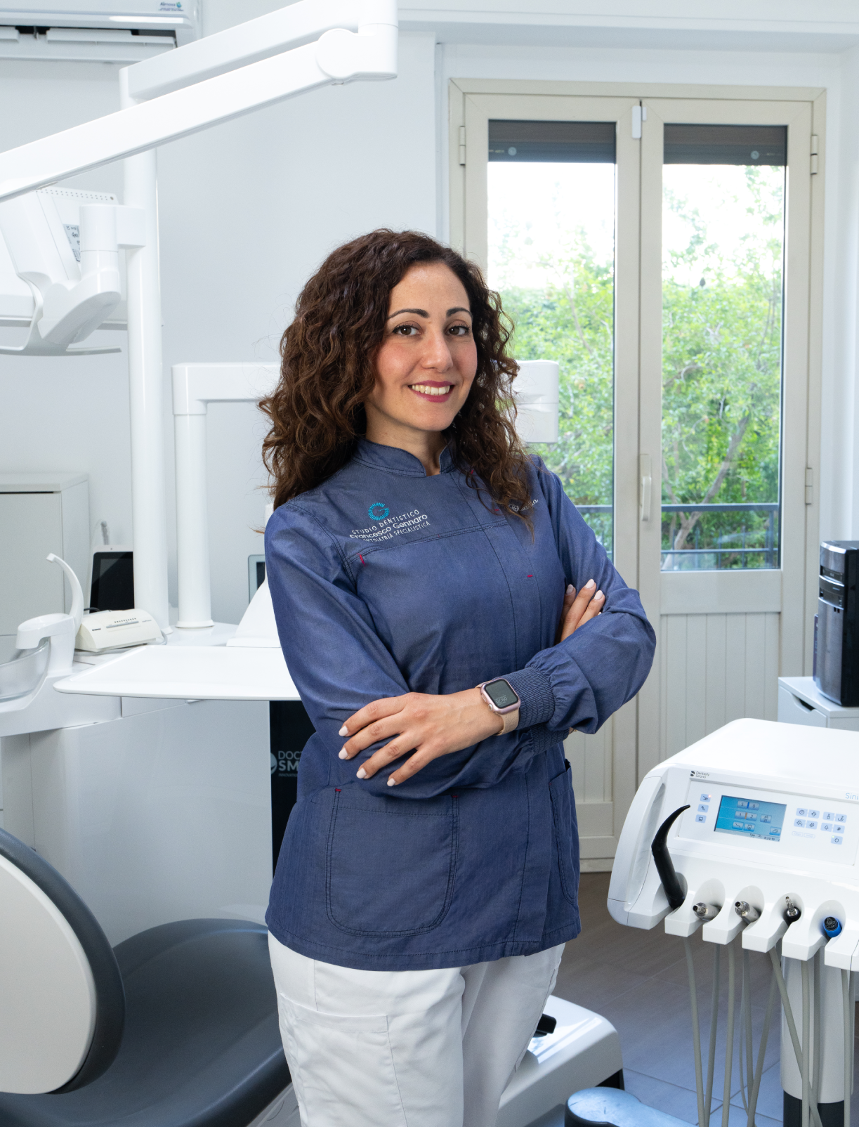 Donna sorridente con una camicia blu in piedi all'aperto accanto a delle piante e una recinzione nera.