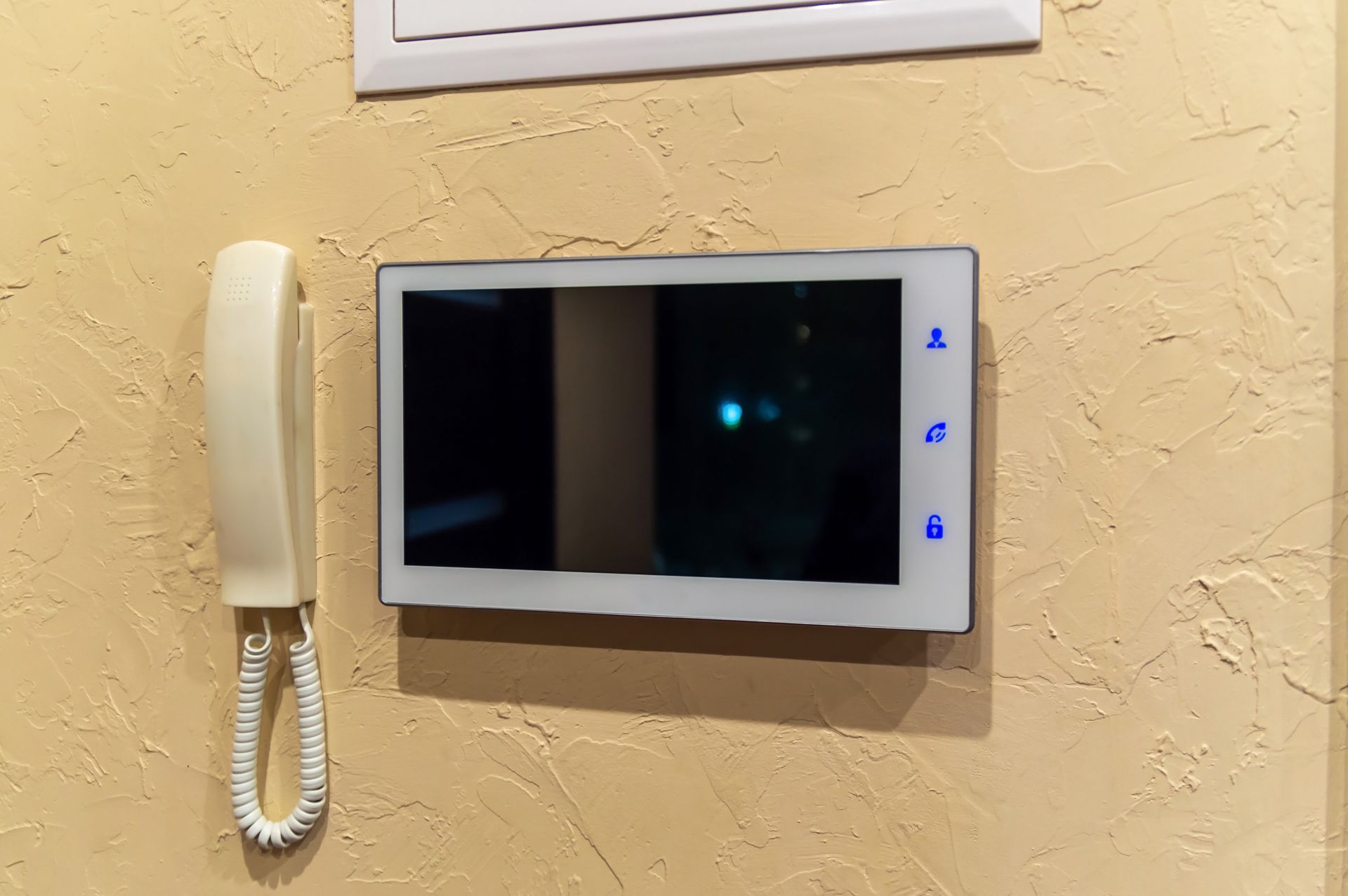 Videocitofono a parete con ricevitore bianco e schermo nero su una parete beige con finitura ruvida.