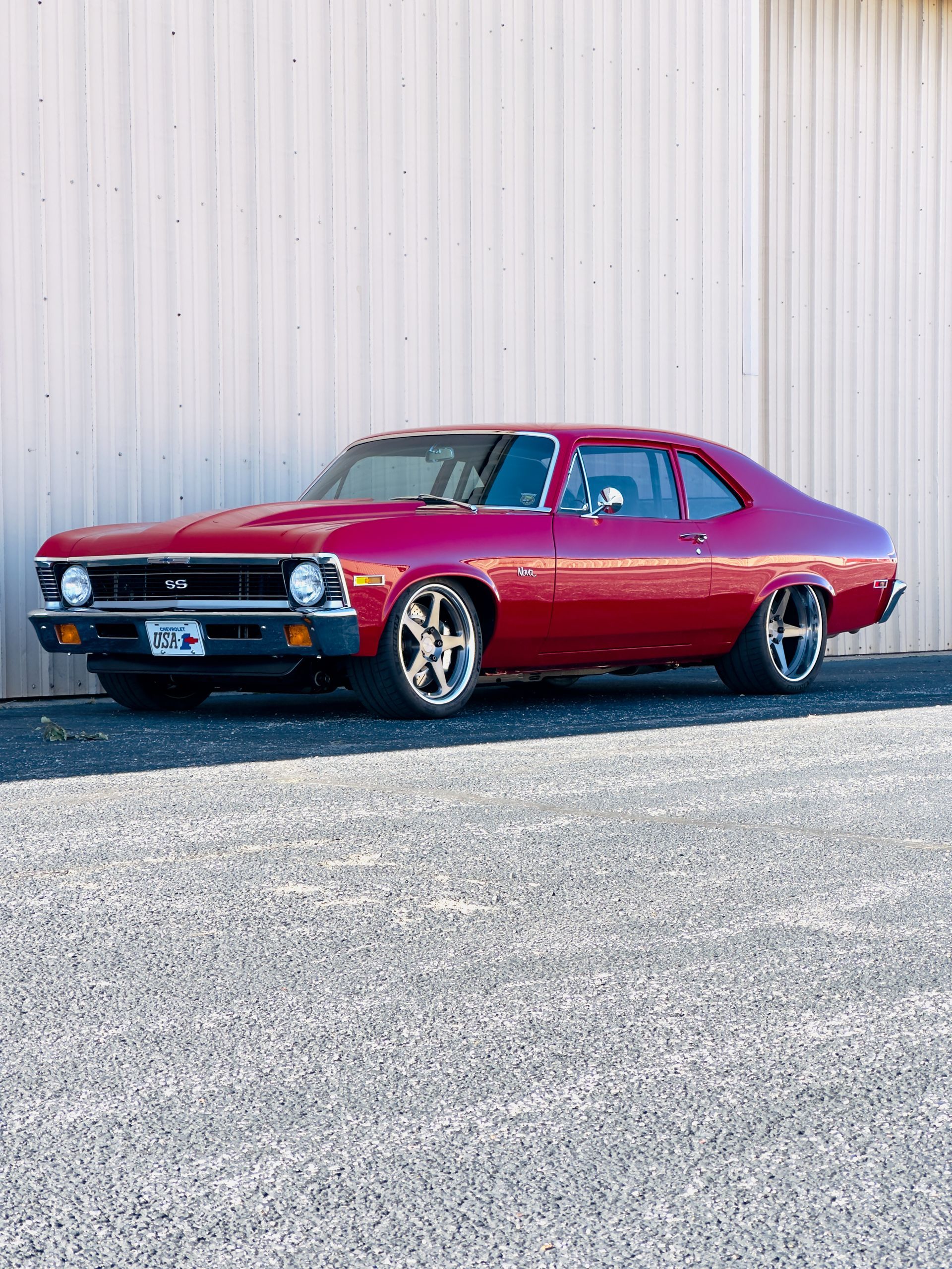 Red classic muscle car parked on gravel in front of a beige metal wall.