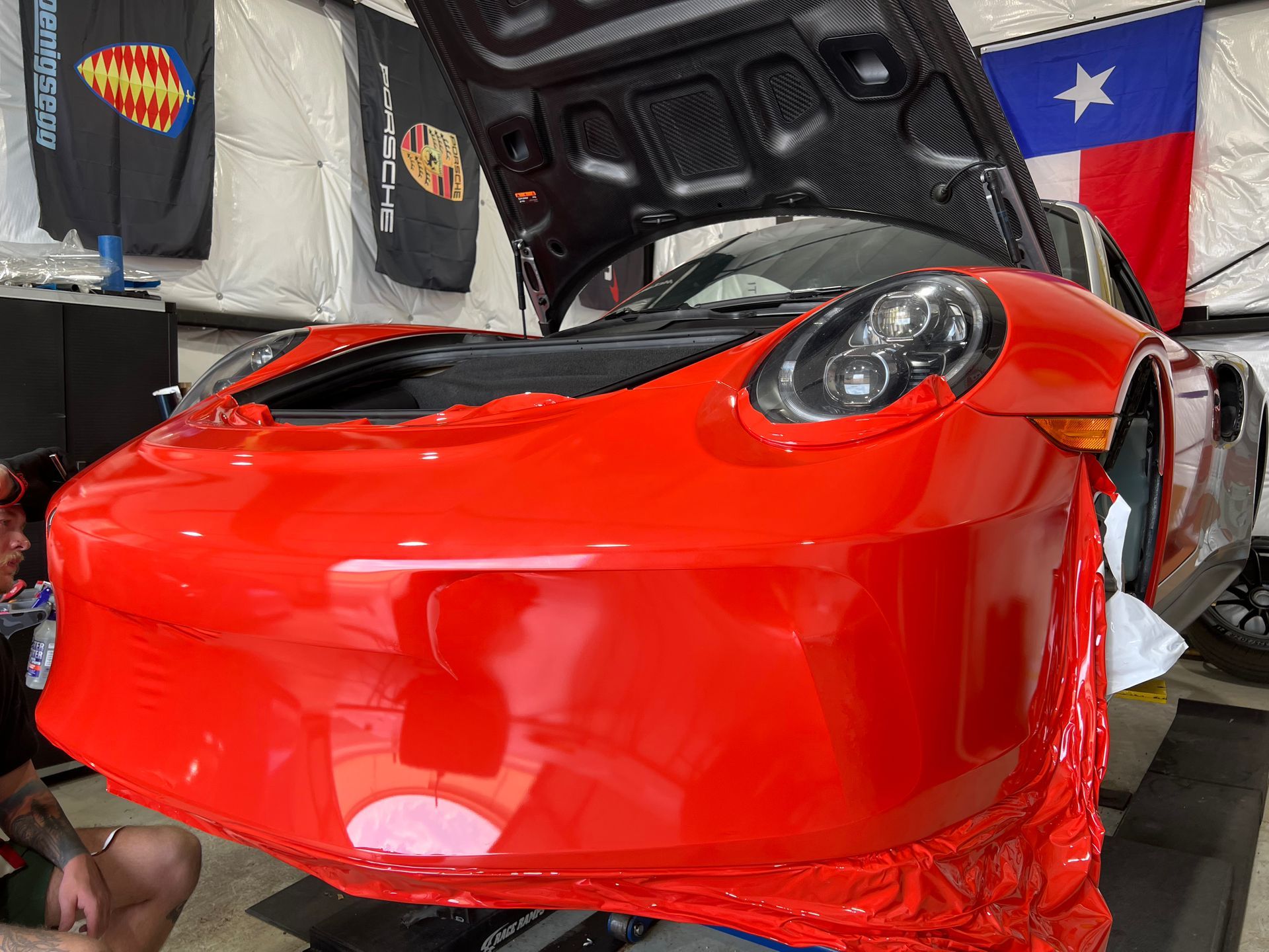 Red sports car in a garage with its hood open, showing a glossy front end and Texas flag in the background