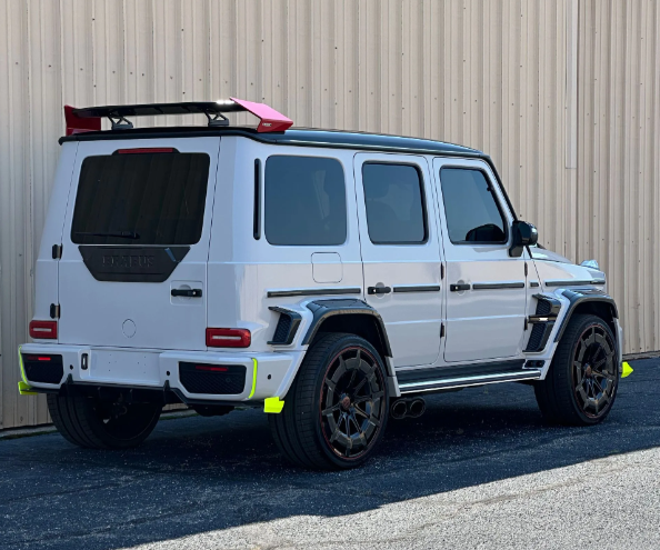 White Mercedes G-Wagon SUV with black trim and roof rack, parked beside a corrugated metal building.