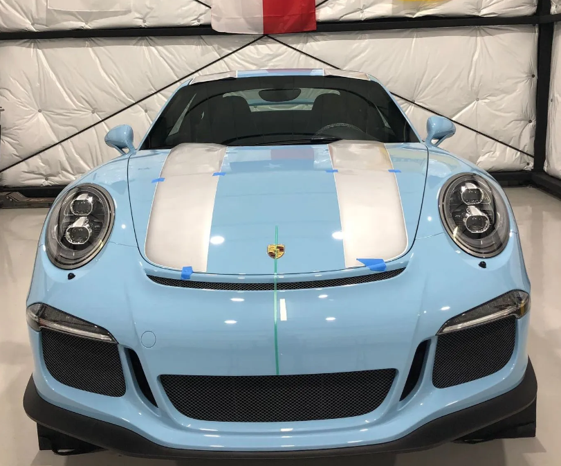 Light blue Porsche sports car with white racing stripes, parked indoors under bright lighting