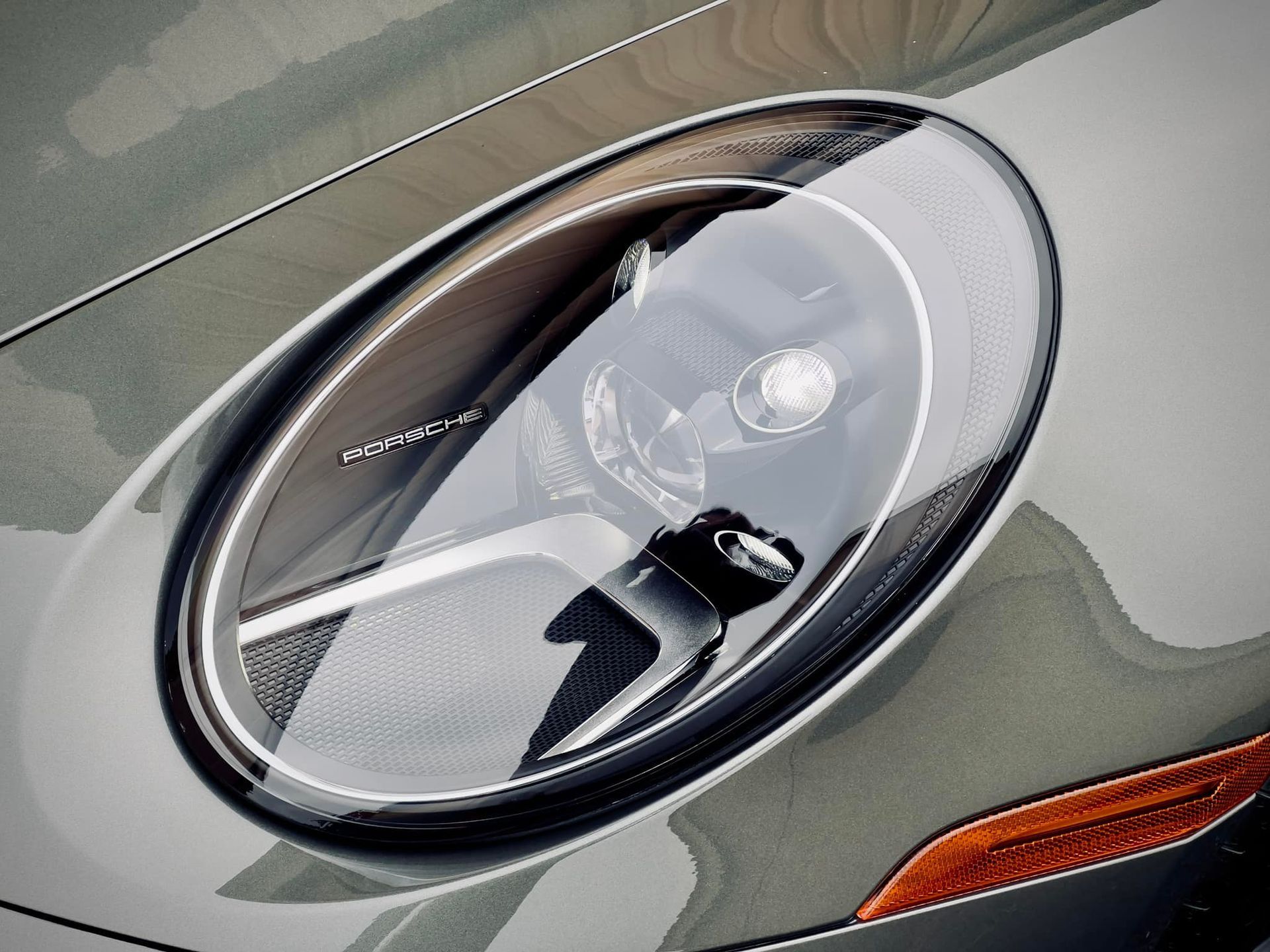 Close-up of a Porsche car headlight, oval shaped, with a reflective lens and dark green metallic paint.