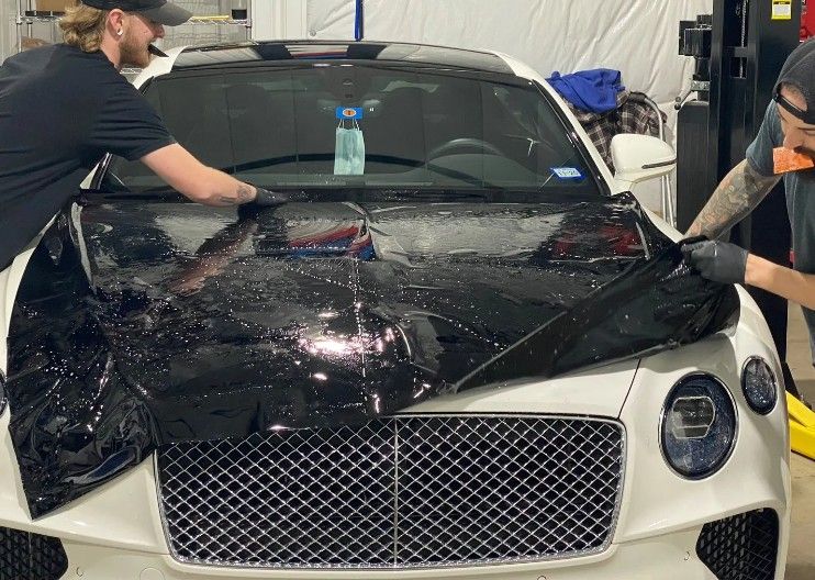 Two people peel clear film off a white luxury car’s hood and windshield in a workshop.