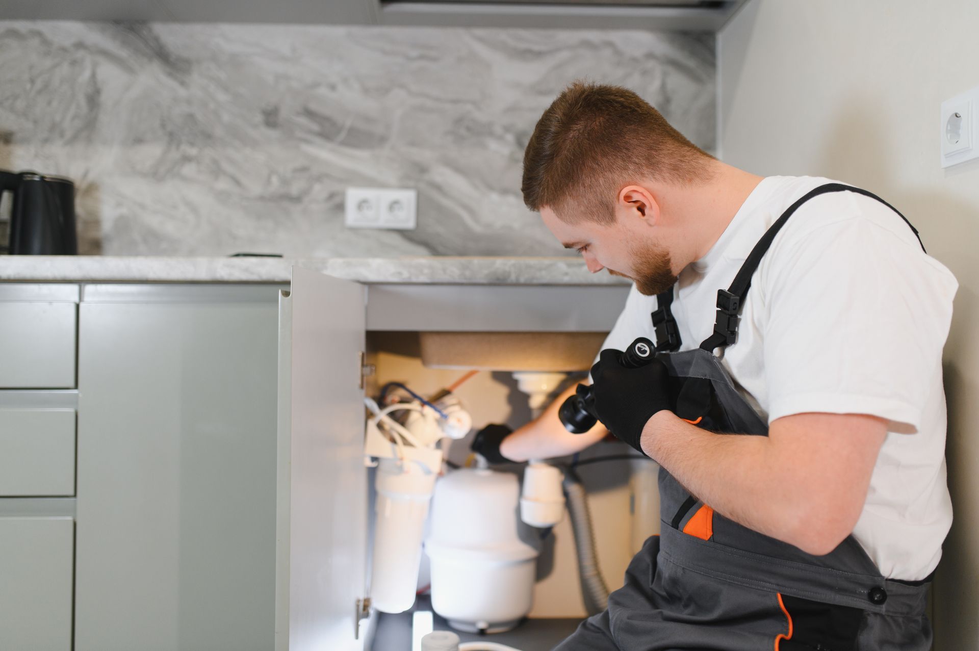Plumber repairing kitchen sink with flashlight.