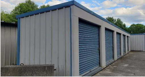 Row of storage units with blue doors, a concrete floor, and a blue-trimmed roof. Trees are visible in the background.