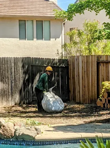 Worker in yellow helmet carrying a large trash bag beside a wooden fence in a backyard.
