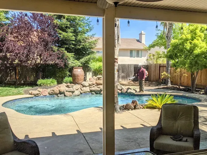 Backyard pool and rock waterfall viewed from a covered patio, with a person standing by the water.