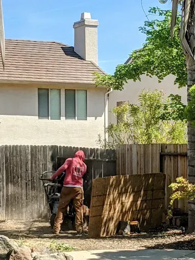 Person in red hoodie handling a wheelbarrow beside a wooden fence in a backyard.