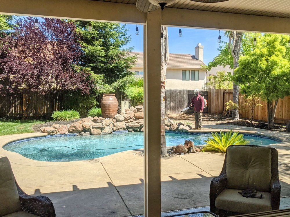Backyard pool with rock waterfall, patio chairs, and a person standing by the water under a covered patio