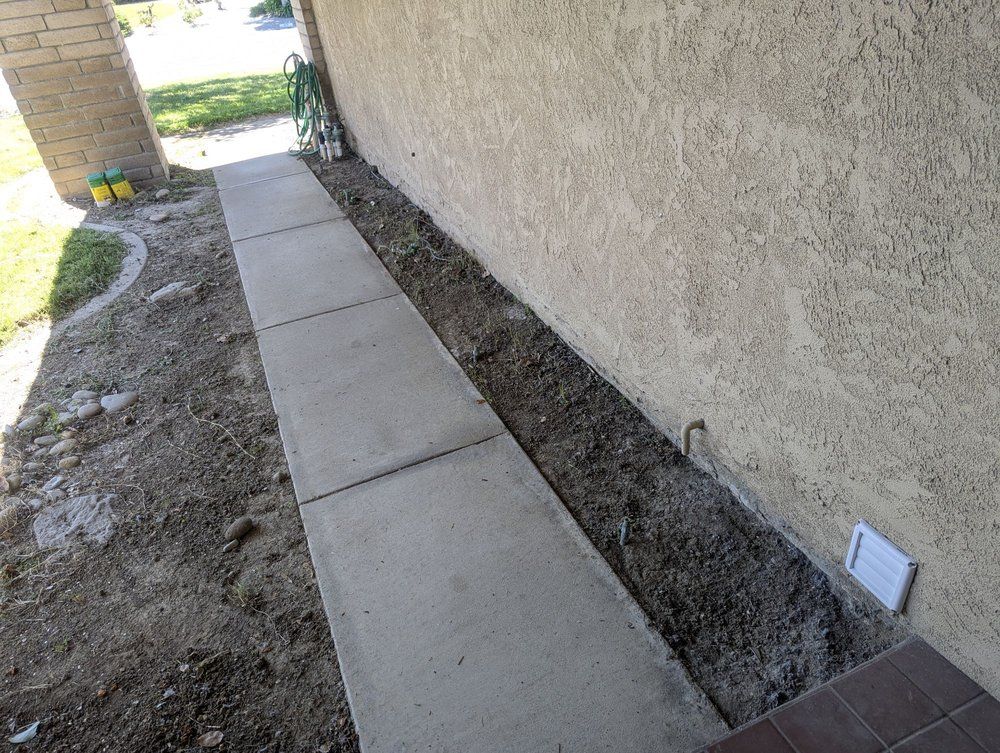 Concrete walkway beside stucco wall, with dirt trench and a small utility box