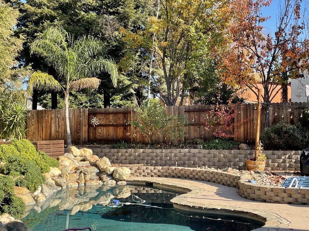 Backyard pool with rock waterfall, wooden fence, and autumn trees