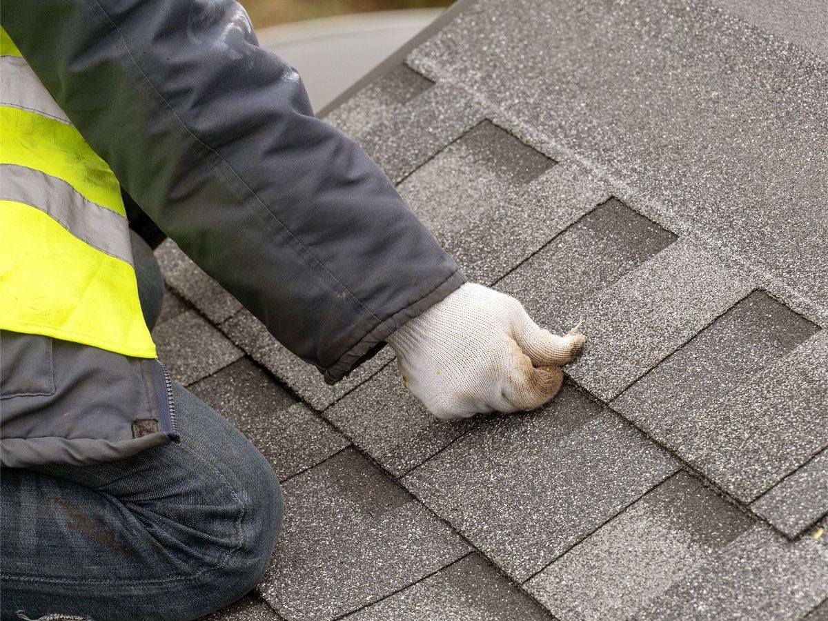 A man is kneeling down on a roof with shingles on it.