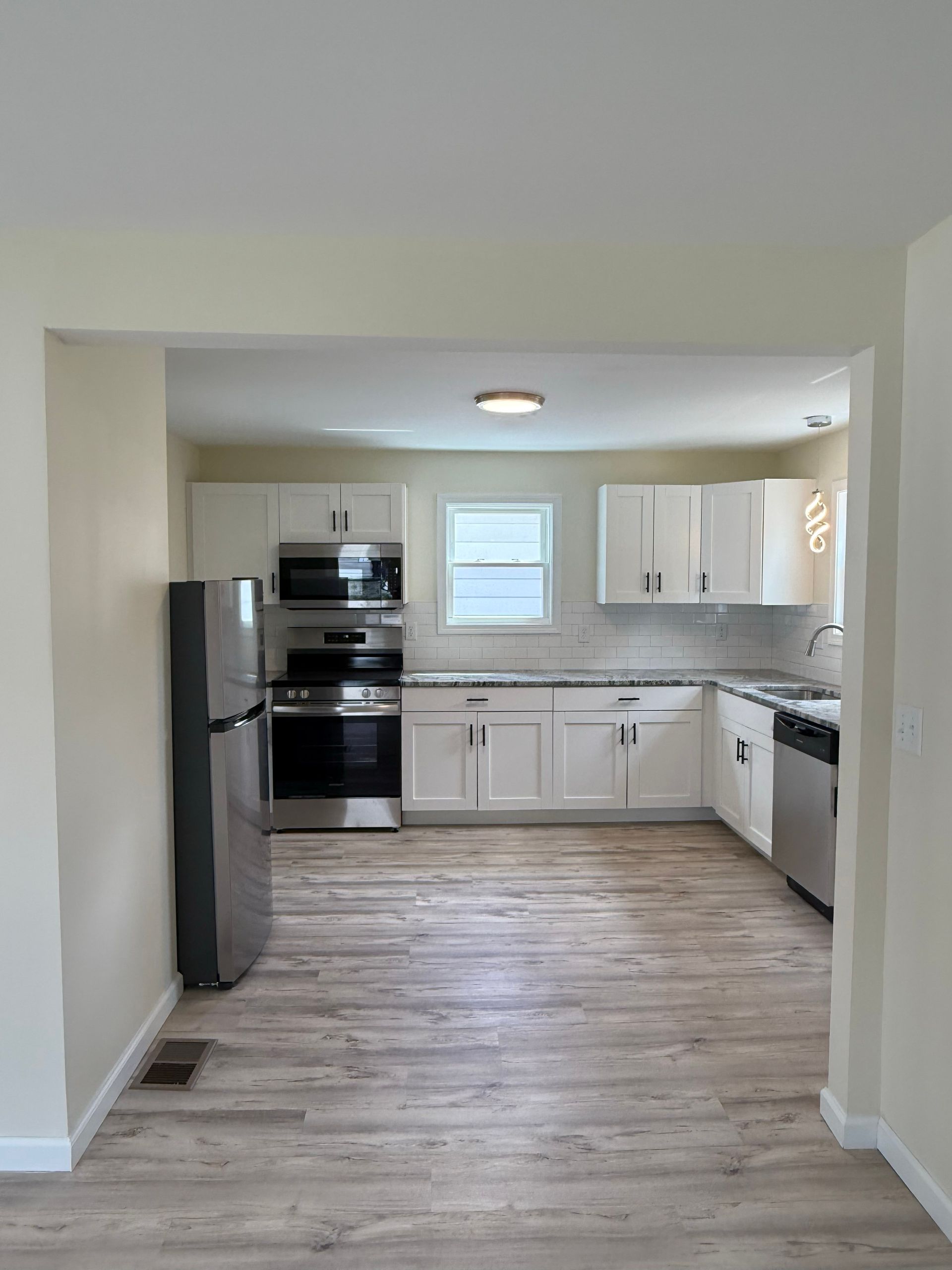 Bright kitchen with white cabinets, stainless steel appliances, and light wood-look flooring. A doorway frames the view.