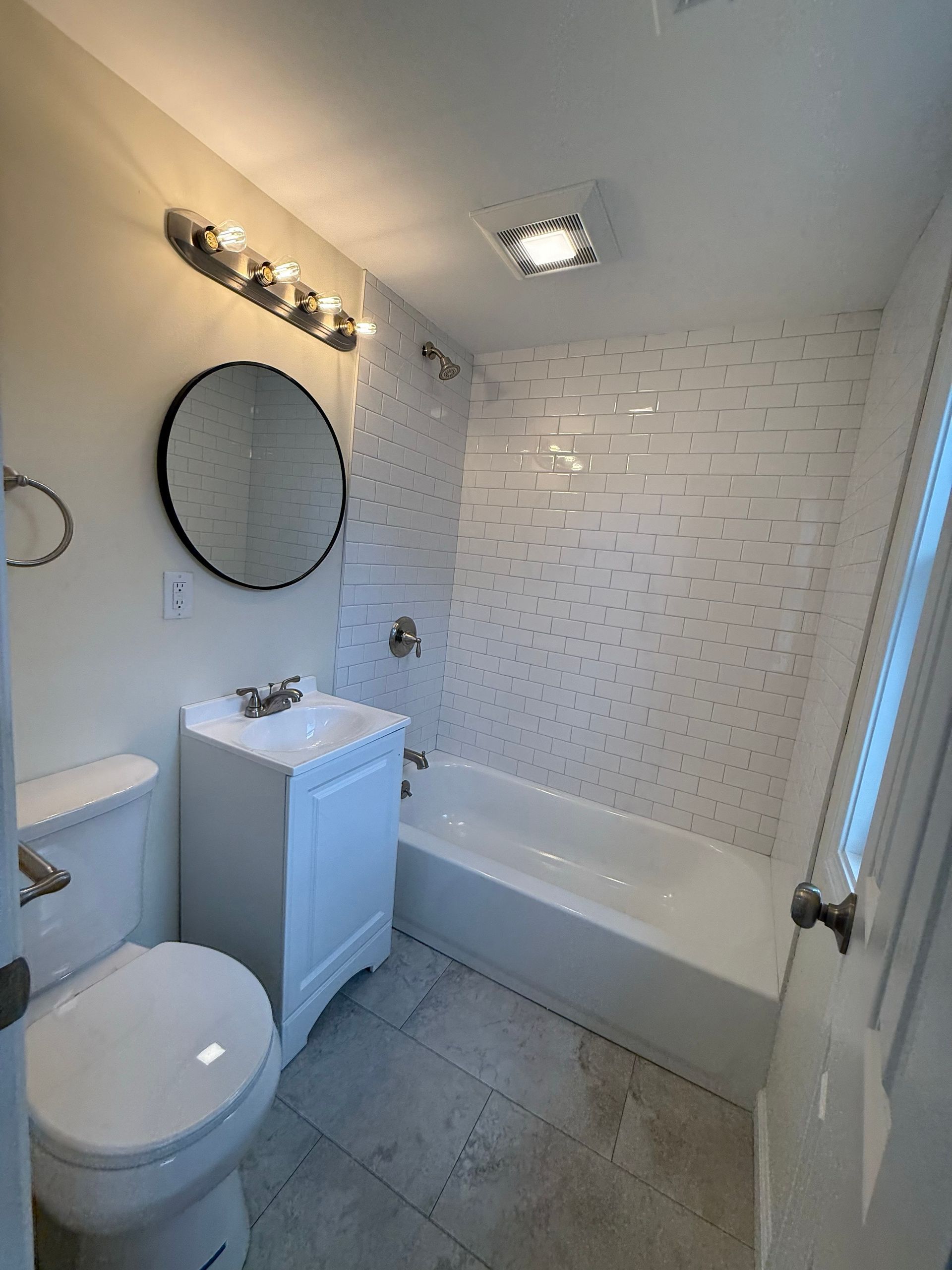 A small, modern bathroom with white tile, a tub, toilet, and a round mirror over a white sink.
