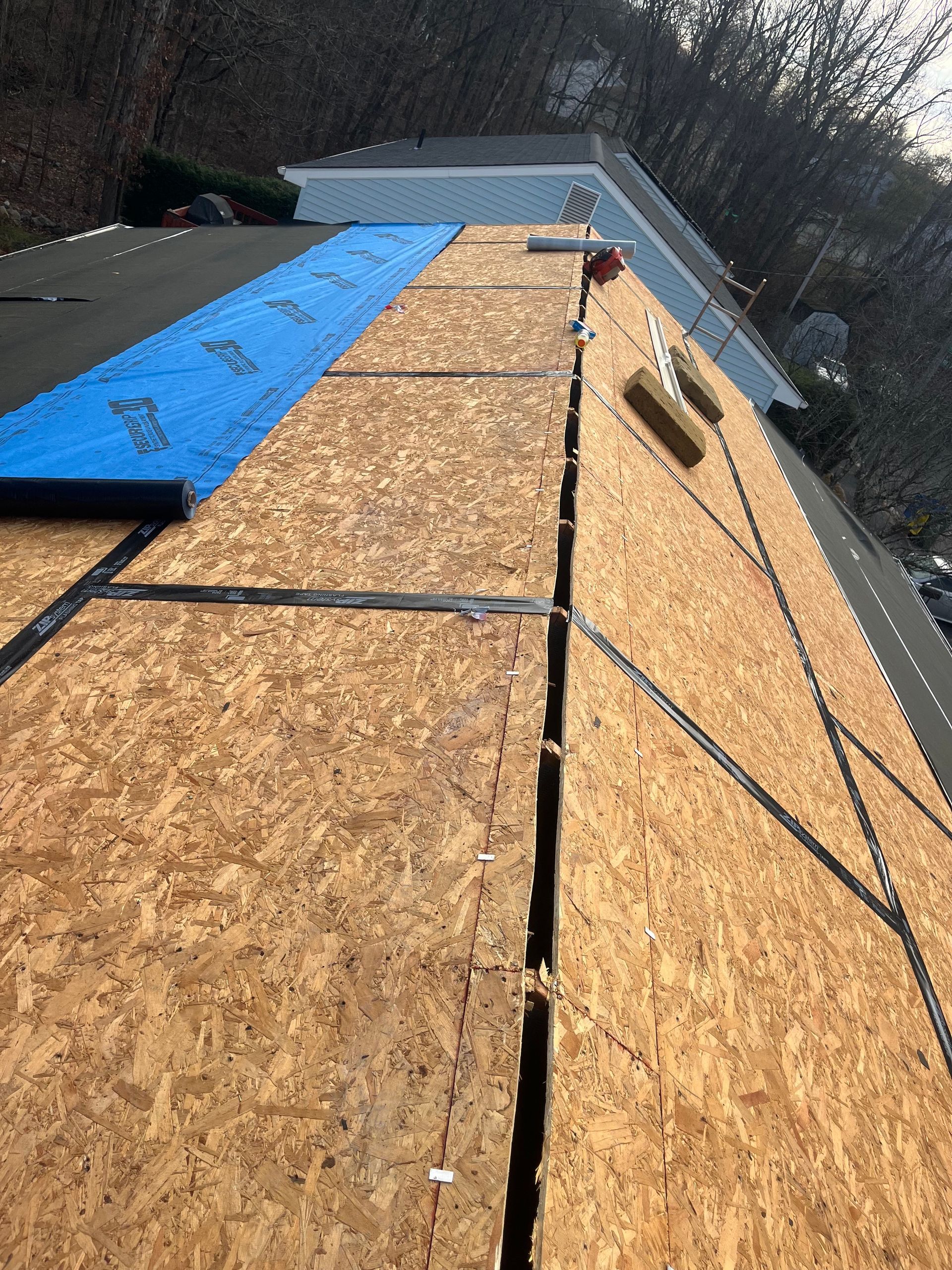Roofing work in progress: wooden boards, blue underlayment, and black sealant on a sloped roof.
