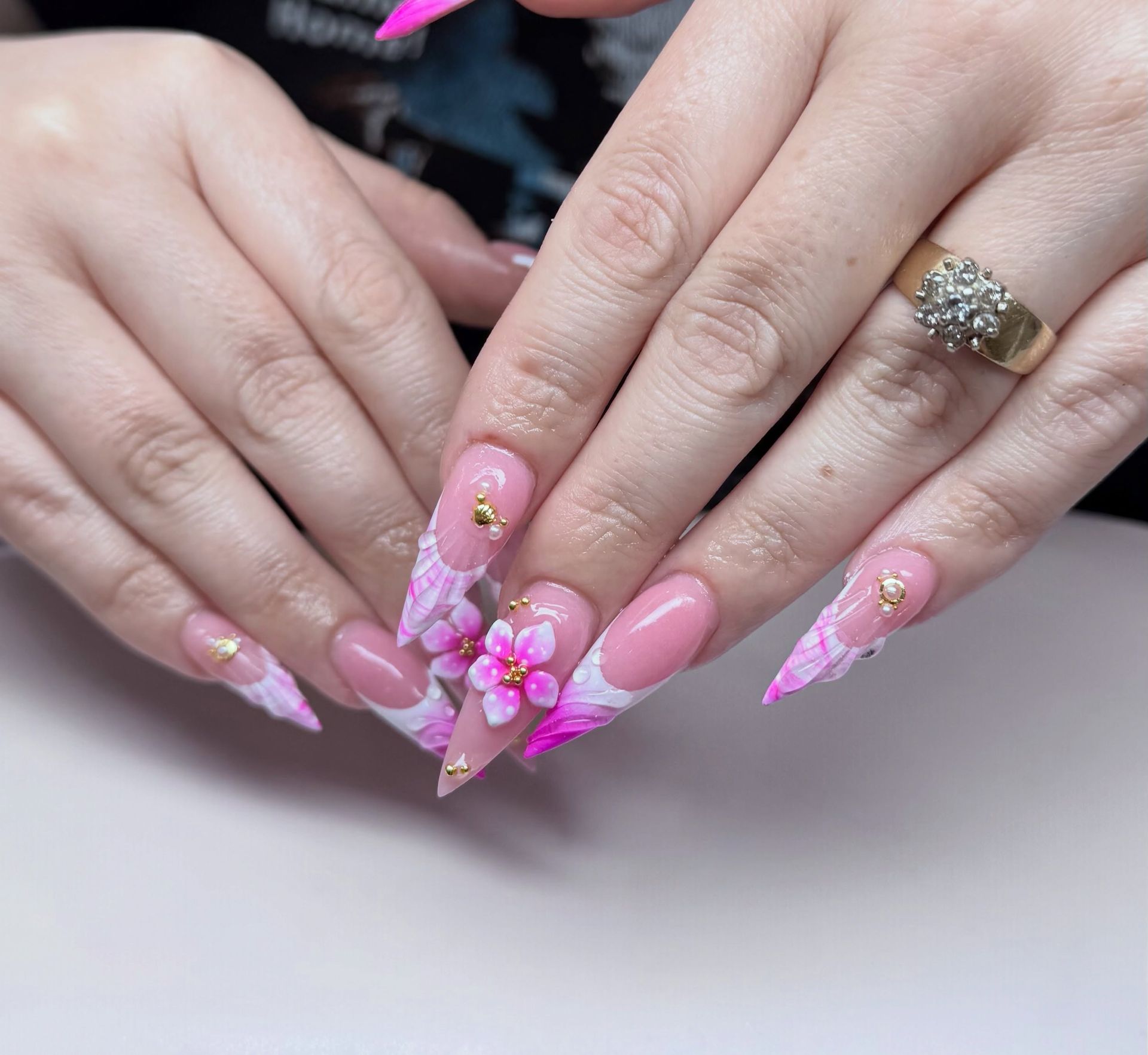 Pink almond-shaped nails with 3D floral accents on both hands, one wearing a gold ring
