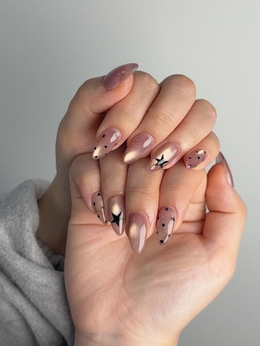 Hands with long nude-pink nails featuring black star accents and gems, resting together on a gray sweater.