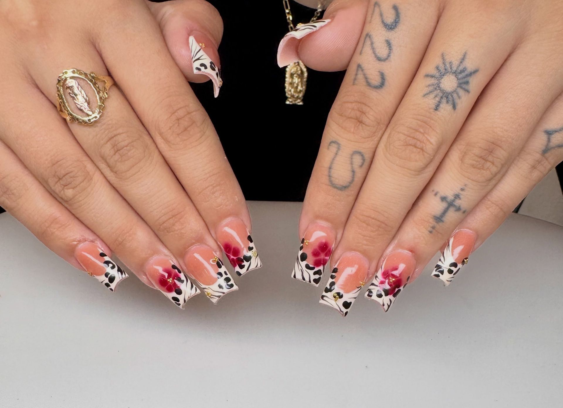 Hands with red and white French-tipped nails, rings, and tattoos resting on a white surface.