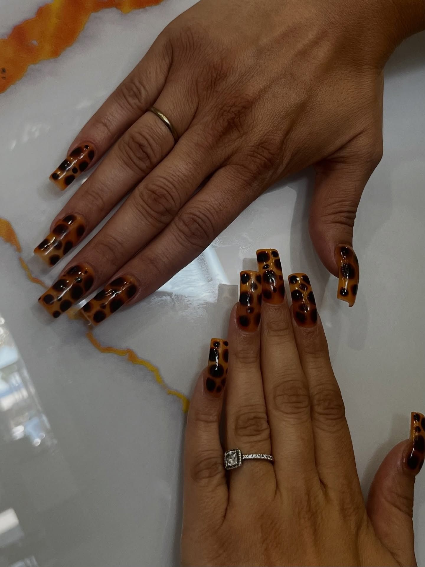 Two hands with long leopard-print nails and rings, displayed on a white marble surface.