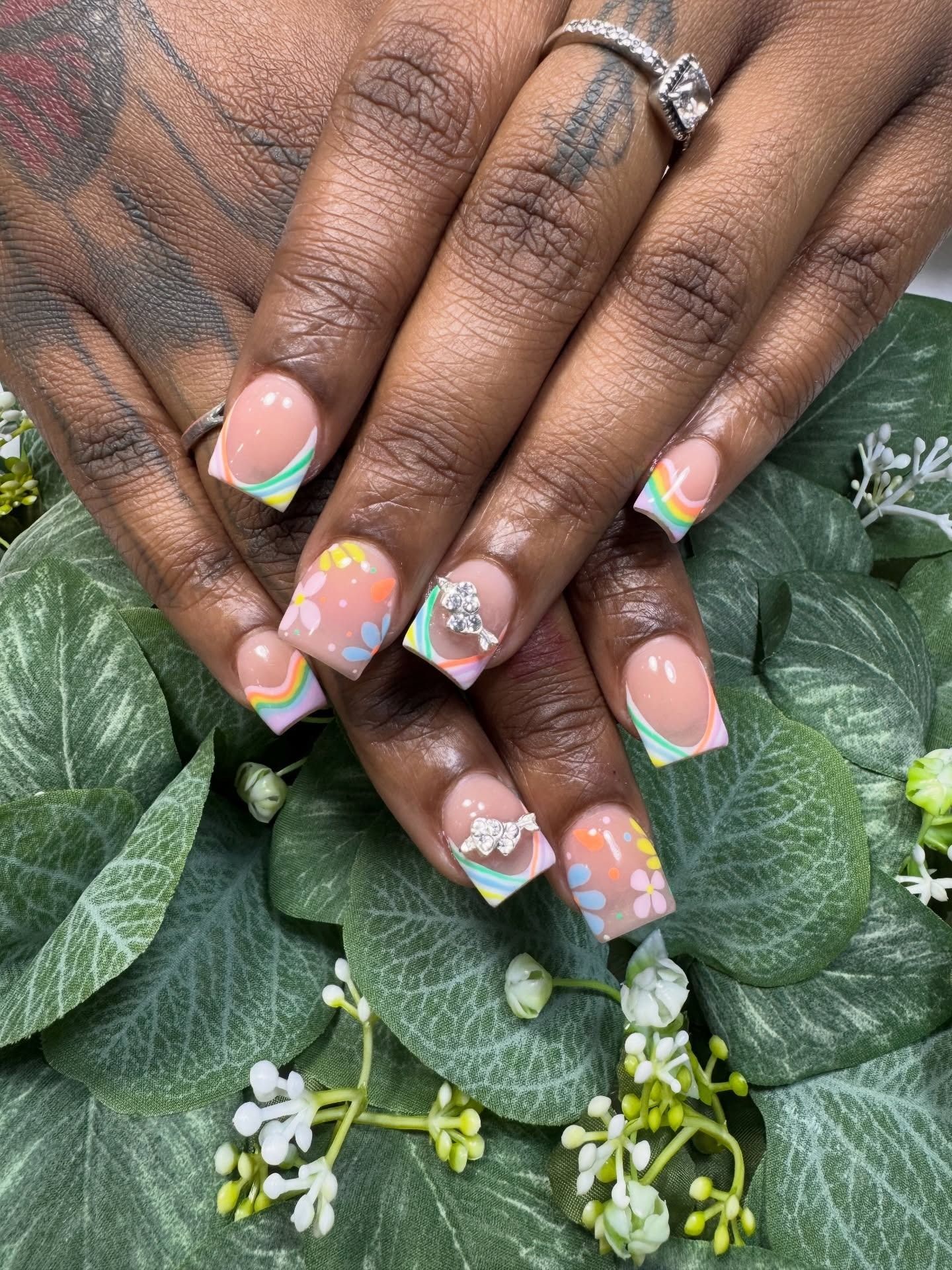 Pastel rainbow nail art on hands resting over green leaves and small white flowers