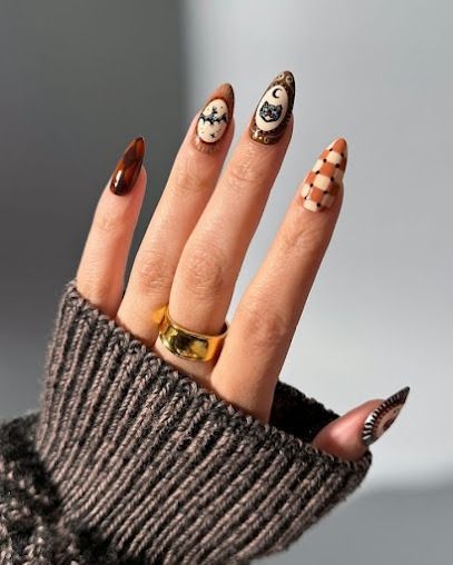 Hand with long nail art in brown, cream, and black, wearing a gold ring and gray knit sleeve.