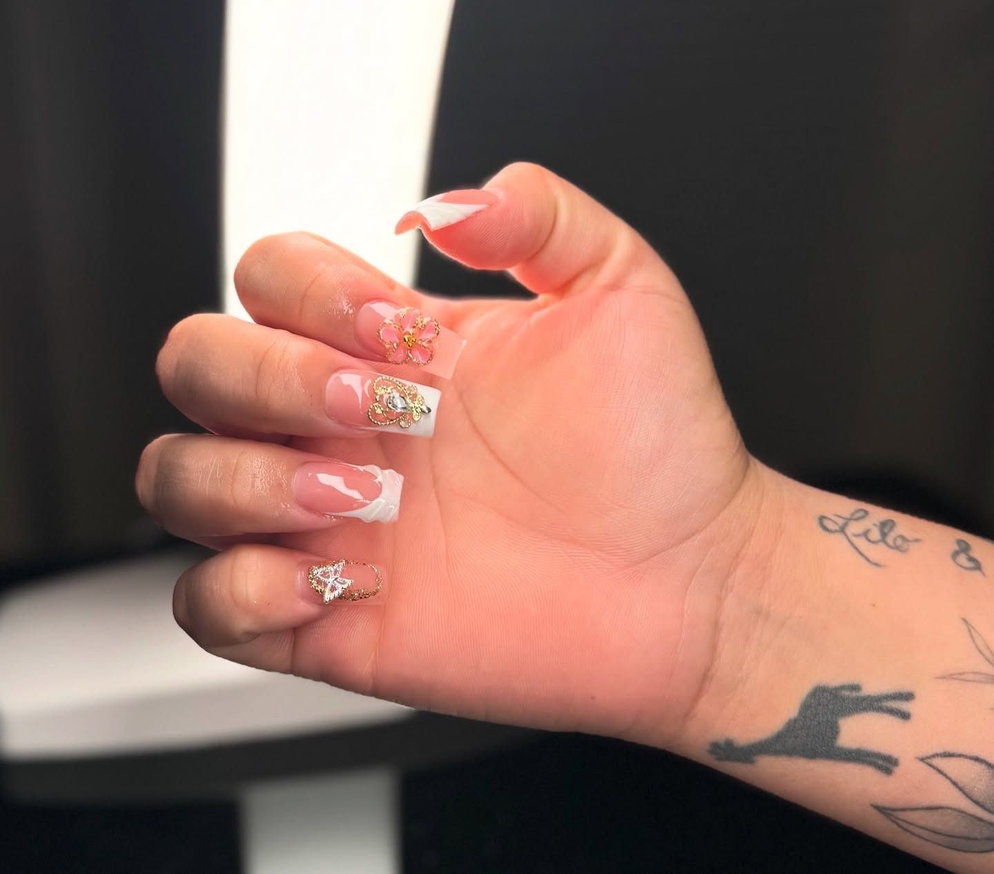 Hand with pink glitter acrylic nails and small charms, shown against a dark background.