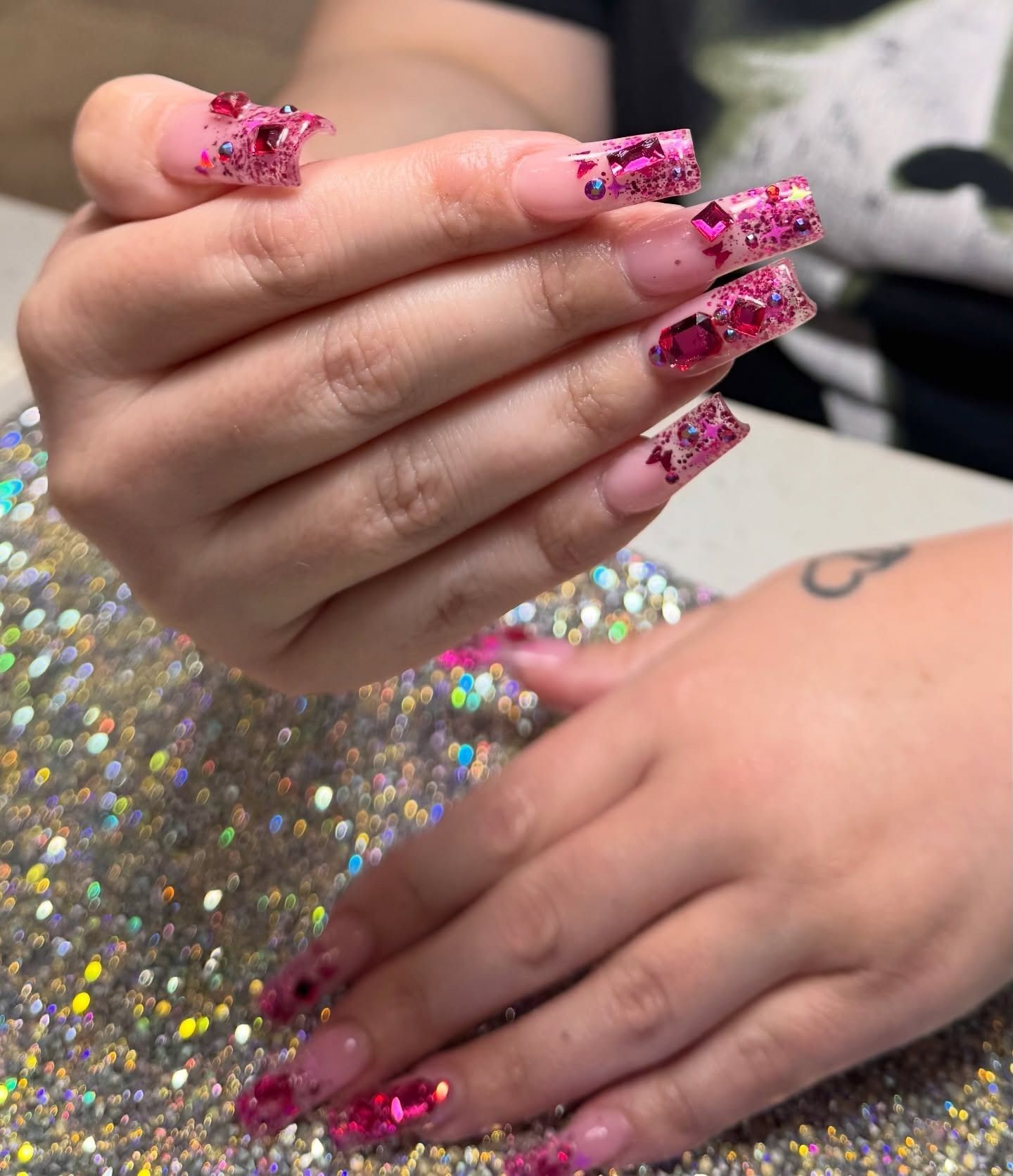Hands with pink glitter nail art over a sparkly silver surface