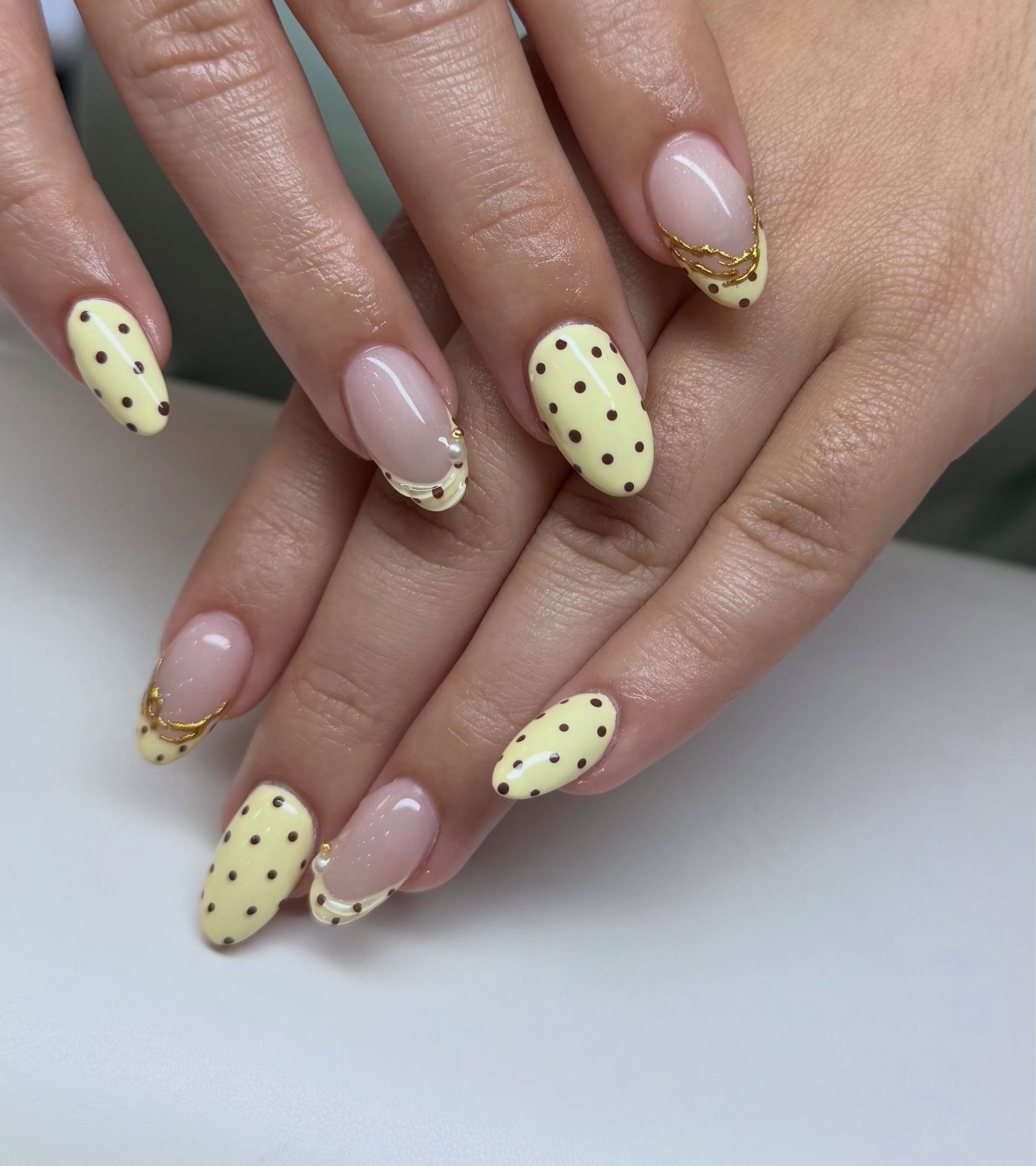 Hands with pastel yellow polka-dot almond nails and gold glitter French tips on a white surface