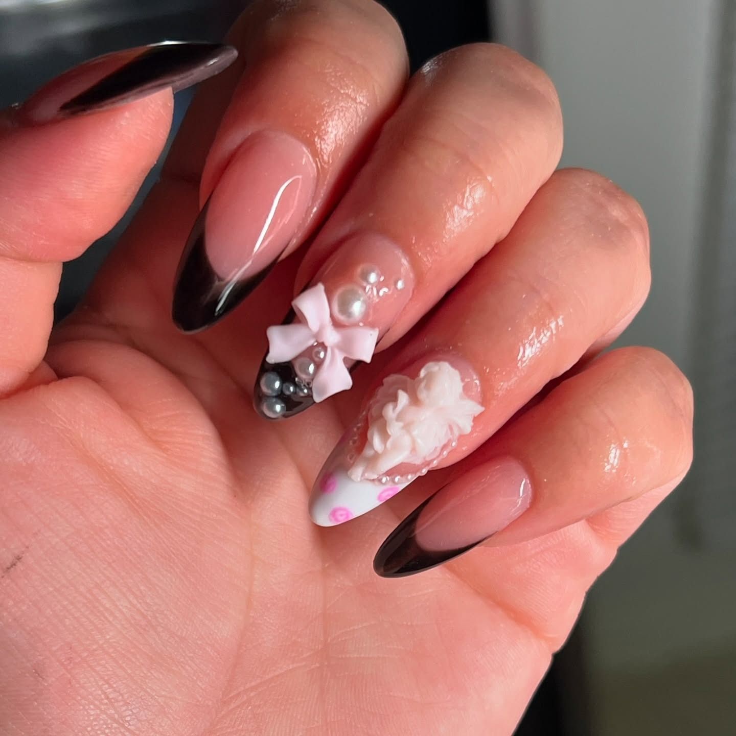 Close-up of manicured hand with black French tips and 3D white-pink nail art, holding a tiny pearl charm.