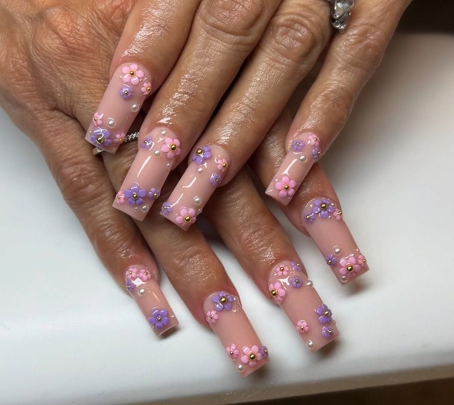 Hands with long clear nails decorated with pink and purple flower nail art on a white surface
